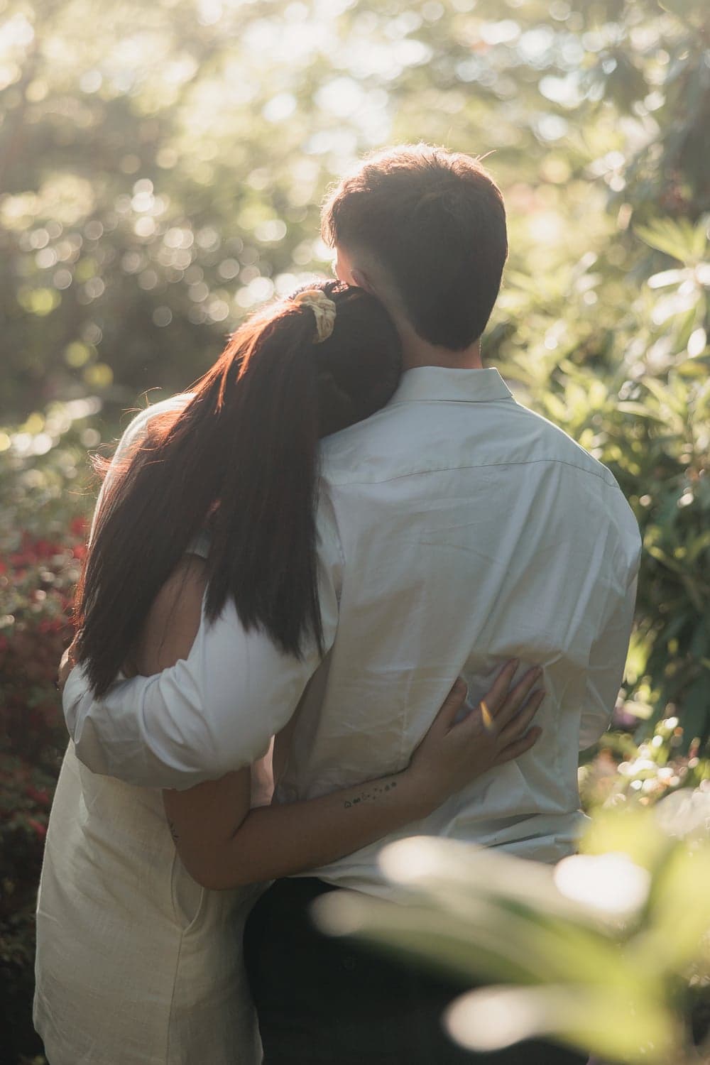 Couple embracing from behind surrounded by sunlit garden foliage during a Canberra engagement shoot.