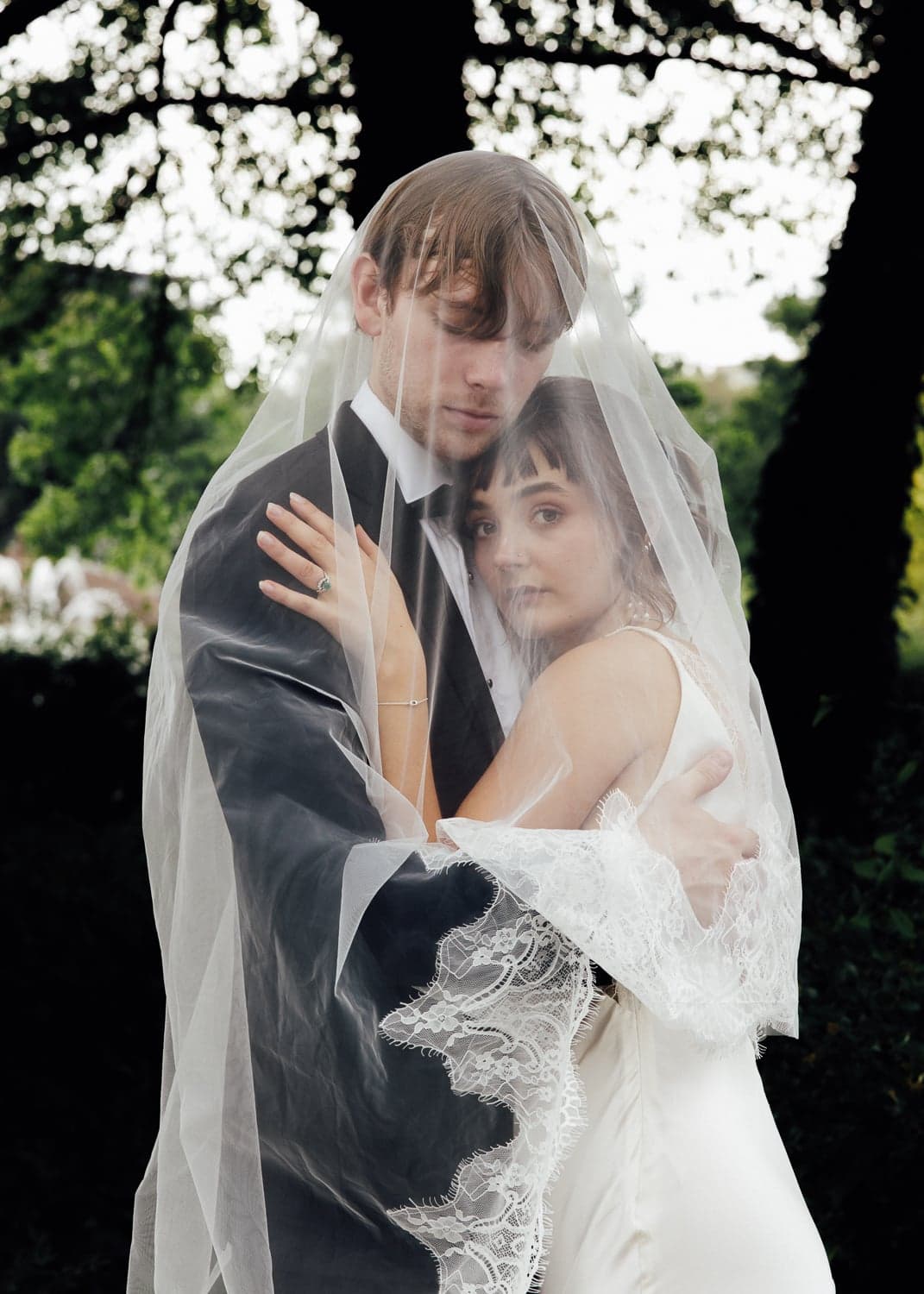Wrapped in a world entirely their own, the couple shelters beneath a lace-edged veil, their closeness soft and unhurried against a backdrop of dappled green. There is a quiet intimacy here that feels like a secret held between two people.