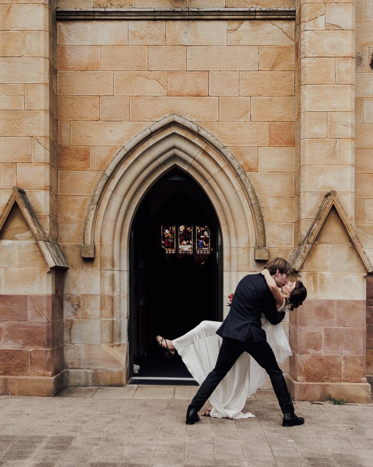 Groom dipping bride in a kiss beneath the arched doorway of St John's Anglican Church, Canberra.
