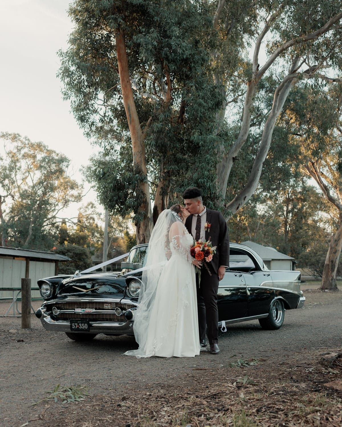 Bride and groom kissing in front of a vintage black car under eucalyptus trees, wedding photography in Canberra.